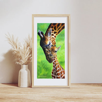 Giraffes - Baby Giraffe Calf and Mom Art Print: A tender wildlife illustration showing a mother giraffe bending her long neck to nuzzle her baby against a vibrant green background, displayed in a light wooden frame with white matting beside a vase of pampas grass