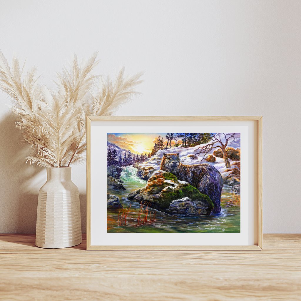 Golden Discovery River Otter art print displayed in wooden frame, showing playful otter on snowy riverbank against vibrant sunset sky. Scenic winter landscape with teal water and evergreen trees. Positioned beside white ceramic vase with pampas grass on wooden surface.