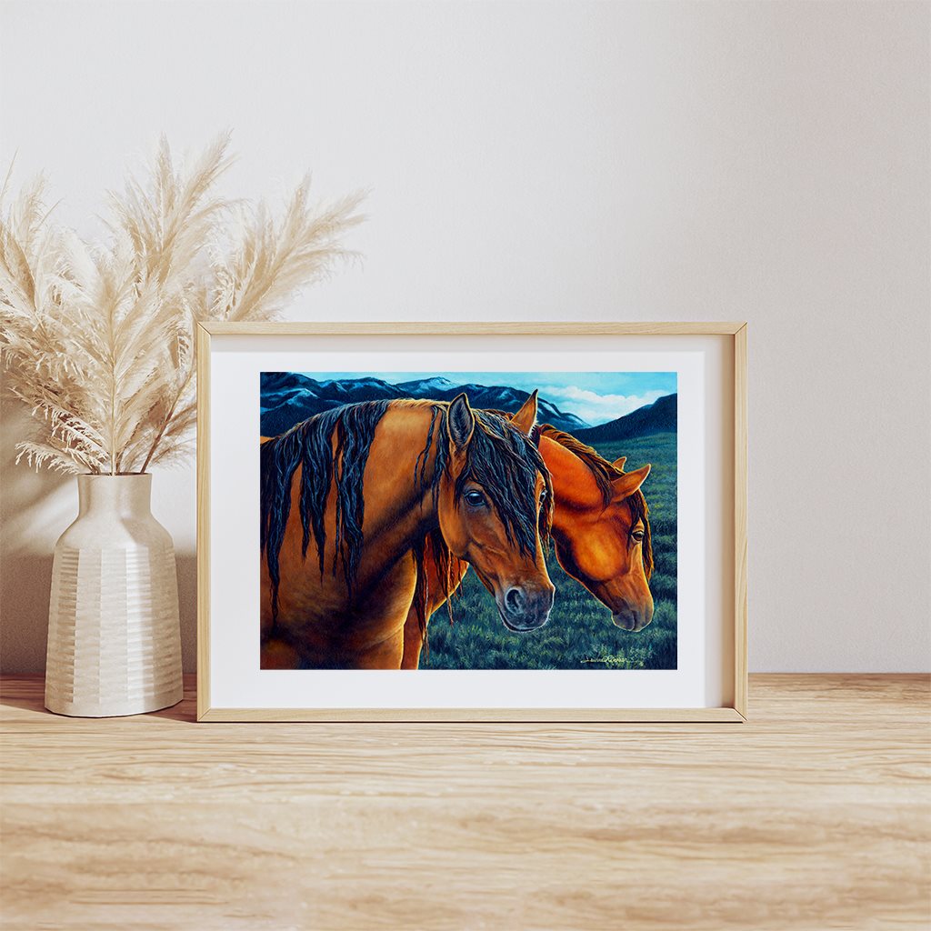 Kiger Mustangs Art Print - Golden Hour features two wild horses with rich chestnut coats standing close together in a mountain meadow at sunset. Their flowing manes catch the warm evening light against a backdrop of blue mountains and grassy terrain.