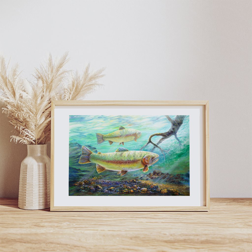 "Rainbow River" art print featuring vibrant rainbow trout swimming in turquoise Alaskan waters, displayed in a light wooden frame next to a cream ceramic vase with pampas grass on a wooden surface. By Jeanne Warren